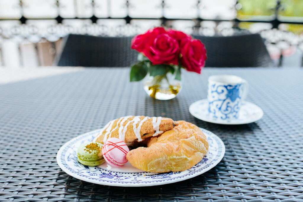 A table on the second floor, New Orleans-style balcony of The William hotel in Boerne, TX. Breakfast pastries and a mug of coffee sit atop the table, which is adorned with a centerpiece of pink roses.