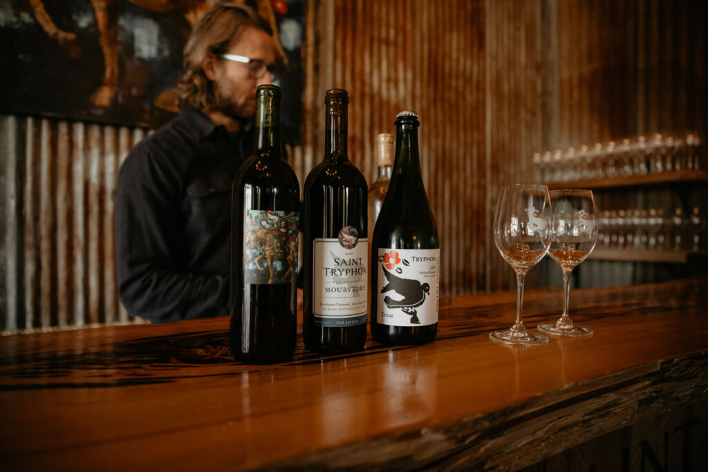 A sommelier behind the counter of a winery. Three bottles of wine and two wine glasses are on the counter.